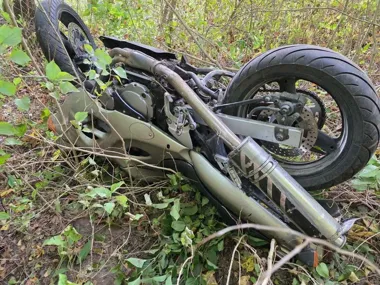 Motocykl leży na boku w zaroślach, częściowo ukryty przez gałęzie i liście.