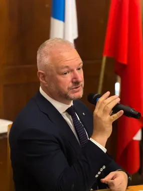 Mężczyzna w garniturze przemawia do mikrofonu podczas oficjalnego spotkania, w tle widoczne flagi.