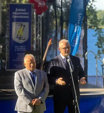 Dwóch mężczyzn w garniturach stoi na scenie nad jeziorem, jeden przemawia do mikrofonu, w tle baner Zakładu Aktywności Zawodowej i flaga.
